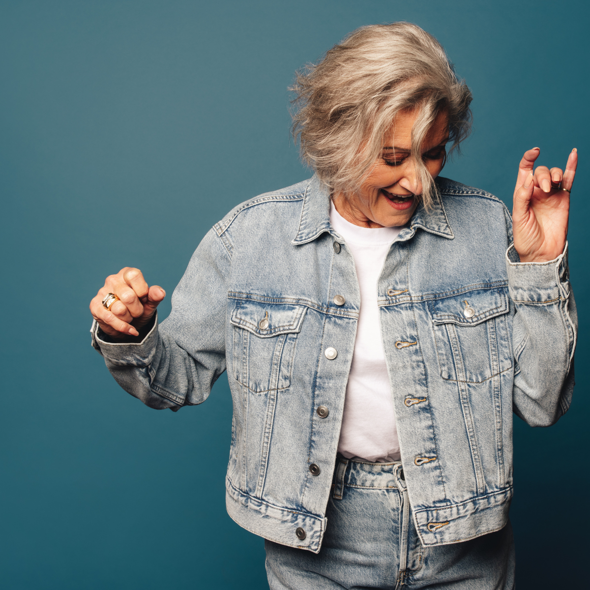 Une femme aux cheveux gris, en veste en jean, danse en souriant devant un fond bleu.