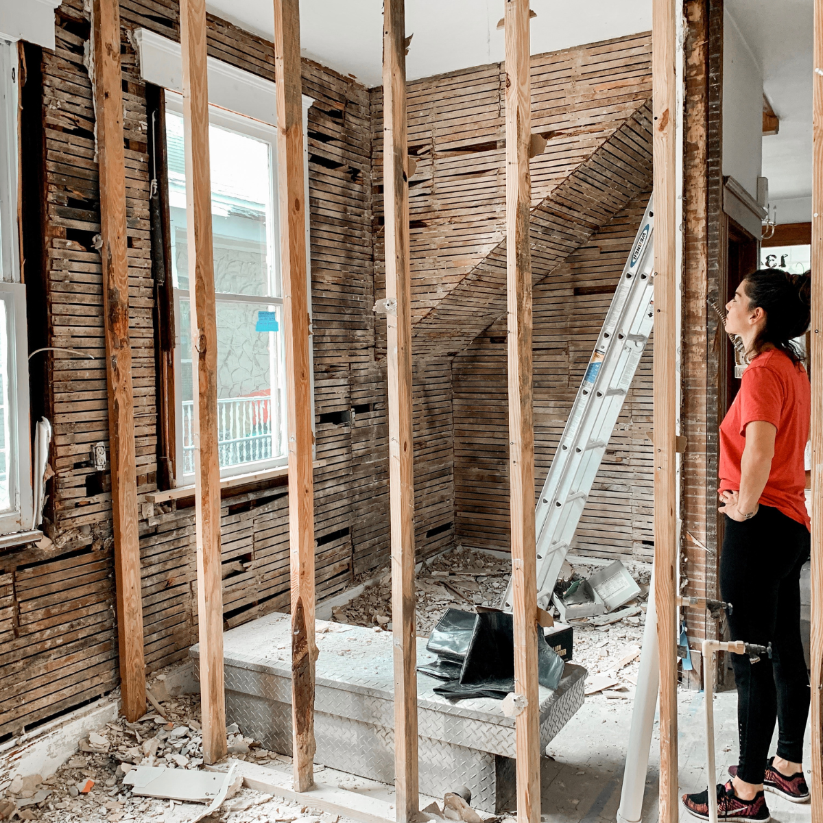 Une femme observe une pièce en cours de travaux, murs ouverts sur les lattes de bois et échelle appuyée au mur.