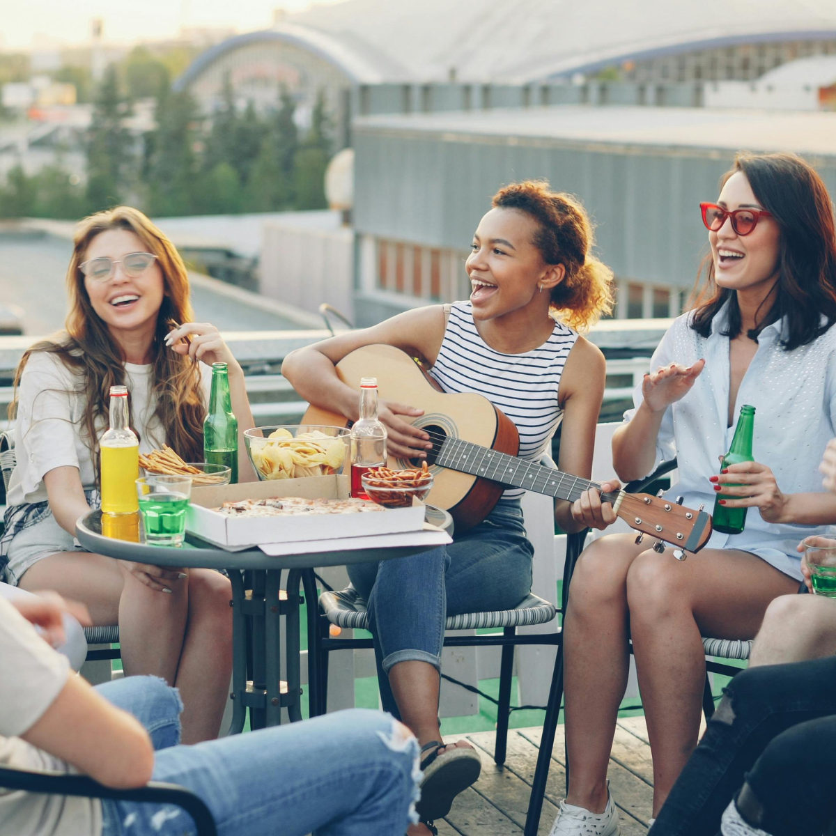 Un groupe de jeunes adultes rit ensemble sur une terrasse en ville, l'un d'eux jouant de la guitare.